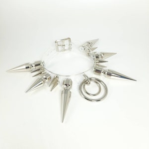 May include: A clear plastic choker with silver spikes and a silver ring attached. The choker is a fashion accessory.