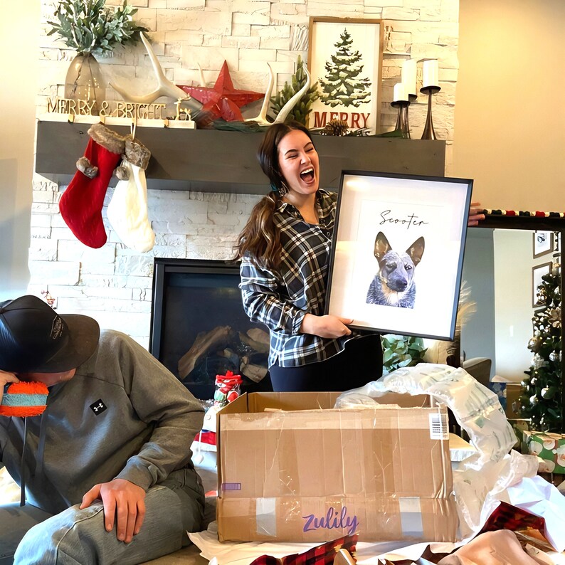 May include: A framed watercolor portrait of a dog named Scooter. The dog is a blue heeler with a black and white face. The frame is black, and the background is white. The portrait is being held by a person in a plaid shirt.