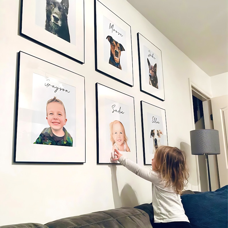 May include: A gallery wall featuring framed portraits of pets and children. Each portrait is in a black frame with a white mat. The portraits include dogs and children with their names written above their images. A child is reaching towards the portraits.
