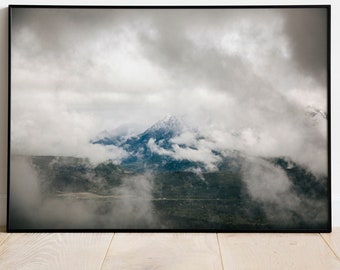 May include: A framed landscape print featuring a mountain range shrouded in thick, gray clouds. The mountain peaks are partially visible, with hints of blue and green in the valleys. The print is set against a white wall and light wood floor.