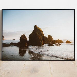 May include: A framed photograph of a coastal scene. The image features large, dark rock formations in the ocean, with waves gently lapping the shore. The sky is a soft blue, and the sunlight creates a warm glow.