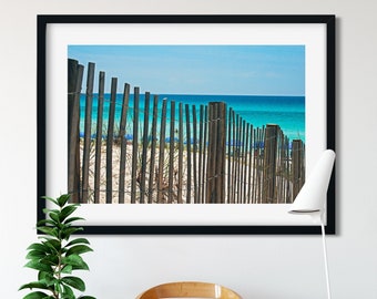 May include: A framed print of a beach scene with a wooden fence, white sand, and turquoise water under a blue sky. The print hangs above a wooden chair with books, a potted plant, and a modern floor lamp.