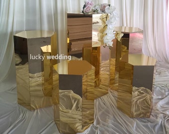 Puede incluir: Conjunto de cinco pedestales hexagonales dorados para exhibir centros de mesa de boda u otros artículos decorativos. Los pedestales están hechos de acrílico espejado y están dispuestos en un patrón escalonado.