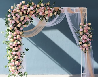 Pode incluir: Um arco de metal branco decorado com uma guirlanda de flores rosa e brancas em cascata. O arco é drapeado com um tecido branco transparente.