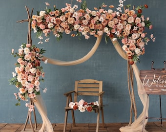 Pode incluir: Um arco de madeira decorado com flores artificiais cor de pêssego e creme. O arco é drapeado com um tecido transparente e uma cadeira de madeira fica na frente dele. Uma placa de madeira com o texto "Bem-vindos ao nosso mundo" está encostada no arco.