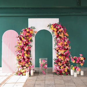 May include: A white backdrop with two arches decorated with pink, orange, and yellow flowers. The backdrop is perfect for a wedding or other special event.