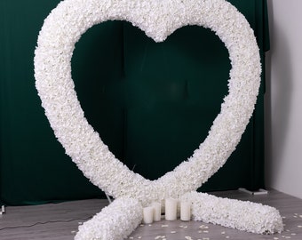 Puede incluir: Un arco floral en forma de corazón blanco con una base blanca, decorado con pétalos de flores blancas en el suelo.