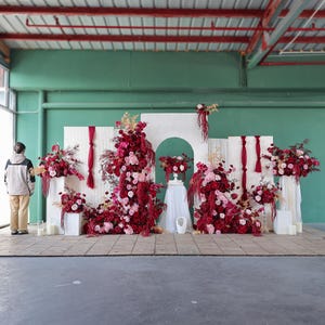 Könnte beinhalten: Eine florale Anordnung mit roten und rosa Blumen, vor weißen Paneelen und einem grünen Hintergrund. Die Präsentation umfasst eine Vielzahl von Blumen und Grün, wodurch eine lebendige und elegante Ästhetik entsteht. Geeignet für Hochzeiten oder besondere Anlässe.
