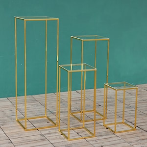 Könnte beinhalten: Satz von vier goldenen Metall-Präsentationsständern mit Glasböden. Die Ständer sind unterschiedlich hoch und in einer Reihe vom höchsten zum niedrigsten angeordnet.