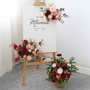 May include: A wooden easel with a white sign that reads "We are Married" with the names "Linjiawei & Guojiaqi" and the date "18.10.16". The easel is decorated with three bouquets of artificial flowers in shades of red, pink, and burgundy.