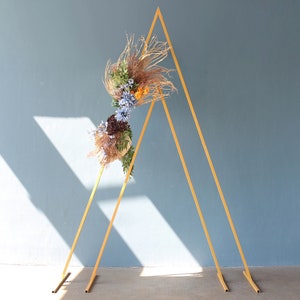Könnte beinhalten: Ein goldfarbener Metallbogen mit einem Blumenarrangement aus getrockneten Blumen und Gräsern. Der Bogen besteht aus zwei dreieckigen Rahmen.