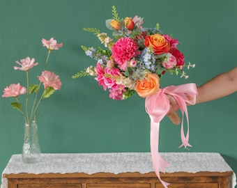 May include: A vibrant bouquet of artificial flowers featuring pink, orange, and red blooms, accented with blue and green foliage. The bouquet is tied with a light pink satin ribbon. A small vase of pink flowers sits on a wooden table.