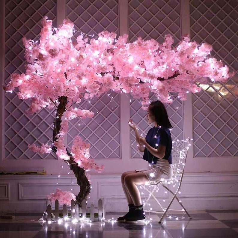 Pu&ograve; includere: Un albero di ciliegio artificiale rosa con luci bianche fatate drappeggiate sui rami. Una persona &egrave; seduta su una sedia bianca di fronte all'albero.