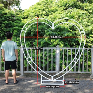 May include: White metal heart-shaped archway with measurements in feet and meters. The archway is 6.5 feet tall and 6.5 feet wide. The base of the archway is 2.2 feet wide and 1.1 feet wide.