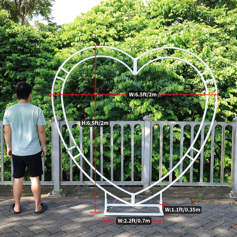 May include: White metal heart-shaped archway with measurements in feet and meters. The archway is 6.5 feet tall and 6.5 feet wide. The base of the archway is 2.2 feet wide and 1.1 feet wide.