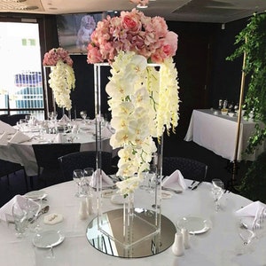 May include: A clear acrylic square vase with a mirrored base, holding a bouquet of pink and white flowers. The flowers are arranged in a cascading style, with the pink flowers on top and the white flowers hanging down. The vase is sitting on a white tablecloth, and there are other floral arrangements in the background.