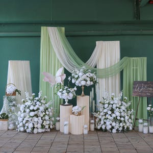 May include: A wedding or event backdrop featuring white floral arrangements, draped fabric in cream and green, and a wooden welcome sign. The scene includes a large paper butterfly and pillar candles, creating an elegant atmosphere.