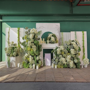 May include: A decorative floral arrangement featuring white and green flowers, candles, and a faux fireplace. The backdrop includes white panels, green accents, and a wooden floor, creating a natural and elegant aesthetic.
