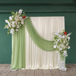 May include: A wedding backdrop featuring ivory curtains, accented with a flowing green fabric. White and green floral arrangements with red accents adorn the backdrop, creating an elegant and romantic setting for ceremonies or photos.