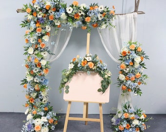 May include: A wedding arch decorated with blue and orange flowers, with a white easel and blank canvas in front of it.