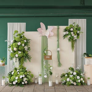 May include: A decorative backdrop with floral arrangements of white and green flowers. A large pink butterfly and a small woven basket are also included. The backdrop is set against a white textured wall with green accents and sheer curtains.