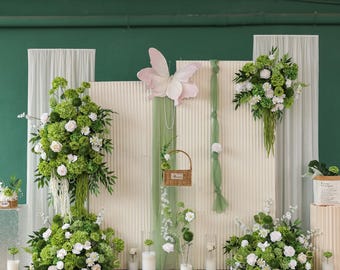 May include: A decorative backdrop featuring floral arrangements with white and green flowers. A large pink butterfly and a small woven basket are also included. The backdrop is set against a white textured wall with green accents and sheer curtains.