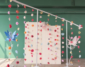 May include: A white metal floral display stand with hanging paper flowers in various colors. The display features two large fairy figures and a backdrop with vertical lines. The flowers are red, pink, orange, and blue.
