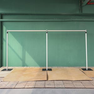 May include: White metal backdrop stand with three adjustable poles. The stand is set up against a green wall and has a brown backdrop on the floor.