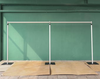 May include: White metal backdrop stand with three adjustable poles. The stand is set up against a green wall and has a brown backdrop on the floor.