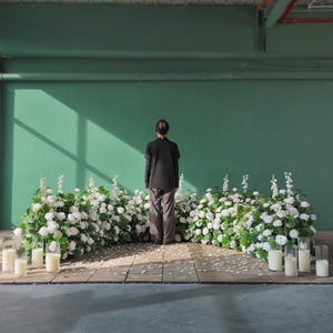 May include: A person stands before a backdrop of white flowers and candles. The person is wearing a black top and gray pants. The backdrop is a green wall with a wooden floor.