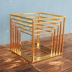 May include: Gold metal tiered stand with four square levels. The stand is made of thin metal bars and has a modern, geometric design.
