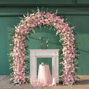 May include: A floral archway, perfect for weddings or events, is decorated with pink and white roses and foliage. The arch frames a white fireplace mantel, with a table draped in pink fabric. A decorative birdcage and a cake are also present.