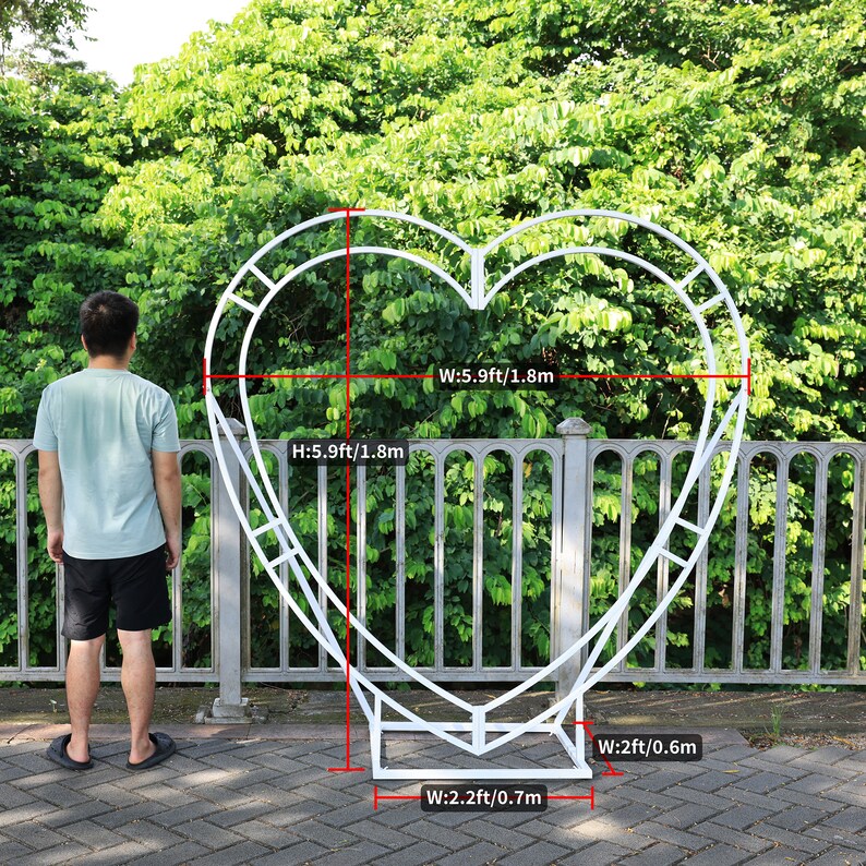 May include: White metal heart-shaped archway with a person standing in front of it. The archway is 5.9 feet tall and 5.9 feet wide. The base of the archway is 2.2 feet wide and 2 feet wide.