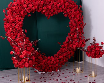 Puede incluir: Un arreglo floral en forma de corazón hecho de rosas rojas, perfecto para una boda o un evento especial. El arreglo se muestra en dos soportes de metal dorado con velas.