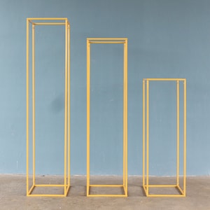 May include: Three gold metal square-shaped stands of different heights. The stands are made of thin metal and have a simple, modern design.