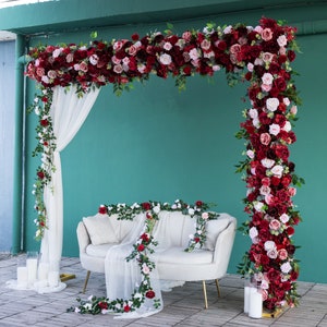 May include: A white couch with a white sheer fabric draped over it, decorated with red and white rose garlands. The couch is set against a green wall with a large floral archway made of red and white roses.