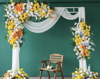 May include: A white fabric backdrop with a wooden frame decorated with yellow and white flower arrangements. A wooden chair sits in front of the backdrop.