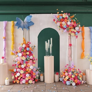 May include: A vibrant floral display featuring a blue butterfly, colourful flowers, and candles. The flowers are arranged in a variety of colours, including pink, orange, and blue. The candles are white and are placed on a beige pedestal.