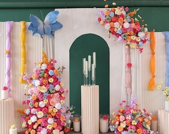 May include: A decorative floral arrangement with a blue butterfly, colorful flowers, and candles. The flowers are arranged in a variety of colors, including pink, orange, and blue. The candles are white and are placed on a beige pedestal.