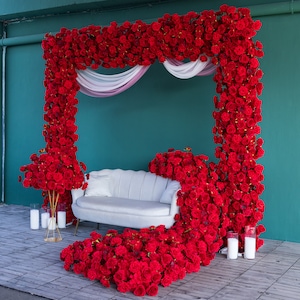 Puede incluir: Un sofá blanco se encuentra frente a un fondo de rosas rojas. Las rosas están dispuestas en forma de marco, con una tela blanca transparente drapeada en la parte superior. El sofá está decorado con almohadas blancas y una manta blanca.