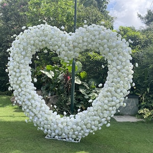 May include: A large white heart-shaped floral arrangement made of white roses. The arrangement is standing on a green lawn in front of a lush green background.