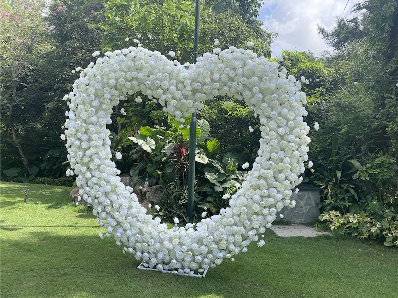 May include: A large white heart-shaped floral arrangement made of white roses. The arrangement is standing on a green lawn in front of a lush green background.
