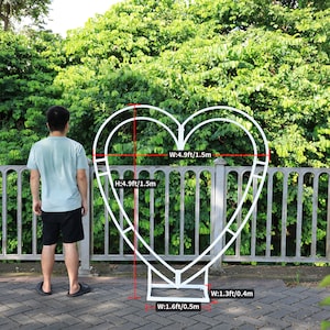 May include: White metal heart-shaped archway with measurements in feet and meters. The archway is 4.9 feet tall and 4.9 feet wide. The base of the archway is 1.6 feet wide and 1.3 feet wide.