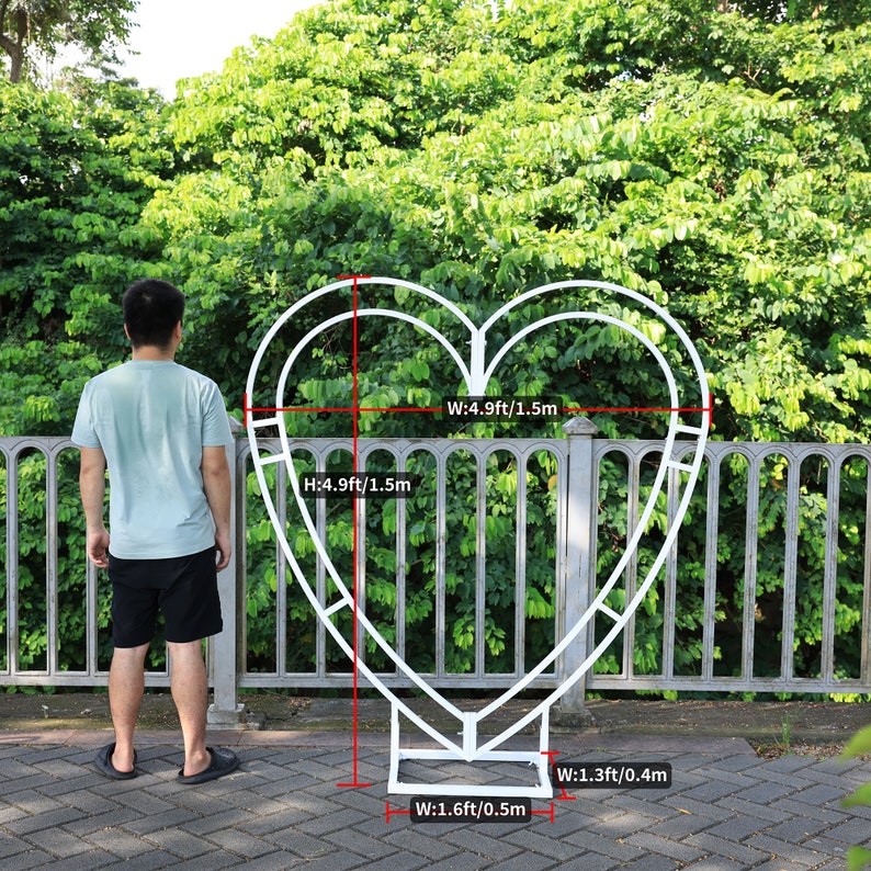 May include: White metal heart-shaped archway with measurements in feet and meters. The archway is 4.9 feet tall and 4.9 feet wide. The base of the archway is 1.6 feet wide and 1.3 feet wide.