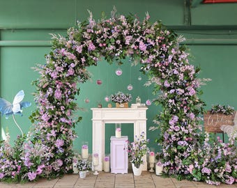 Puede incluir: Un arco floral decorativo adornado con flores rosas y moradas, una chimenea blanca y un letrero de madera con la palabra "Welcome". La escena está ambientada en un suelo de madera con un fondo verde, ideal para bodas o eventos.