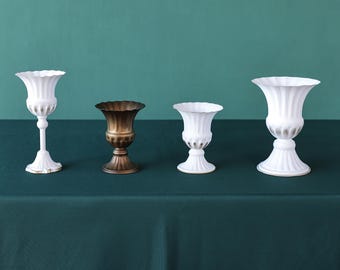 May include: Four decorative vases in a row. Three are white with fluted designs and gold accents, and one is bronze. The vases vary in size and shape, with a pedestal base and ruffled rims. They are arranged on a dark green surface.
