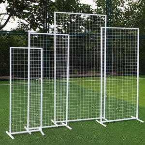 Może przedstawiać: Cztery białe metalowe panele kratowe o różnych rozmiarach, stojące na zielonej trawiastej powierzchni. Panele wykonane są z siatki drucianej i mają prosty, nowoczesny design.