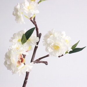 May include: A branch of white artificial cherry blossoms with green leaves. The flowers are clustered together and have a delicate, realistic appearance.