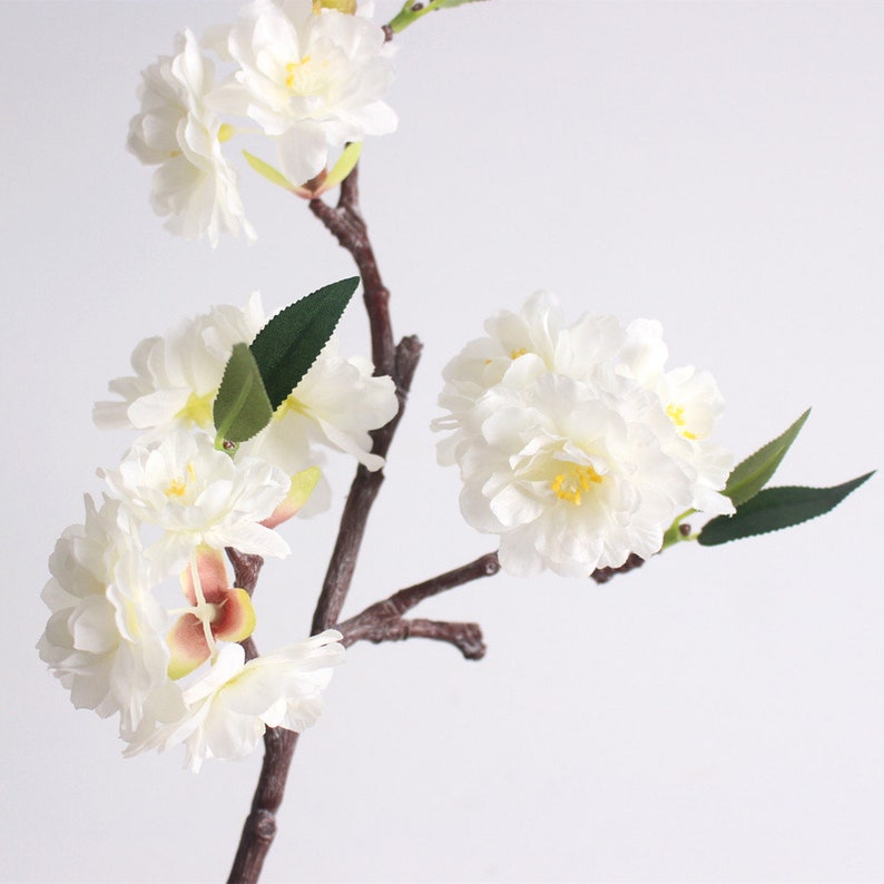 May include: A branch of white artificial cherry blossoms with green leaves. The flowers are clustered together and have a delicate, realistic appearance.