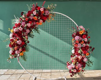 Pode incluir: Um arco circular de metal branco com um padrão de grade, adornado com vibrantes flores vermelhas, rosas e laranjas e vegetação. O arranjo floral é denso e exuberante, criando um belo cenário para eventos.